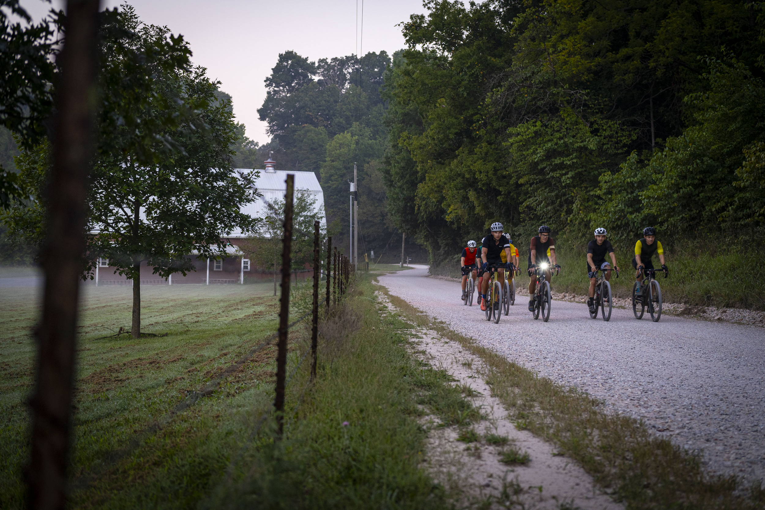 Gravel cyclists ride their bikes past a red barn.