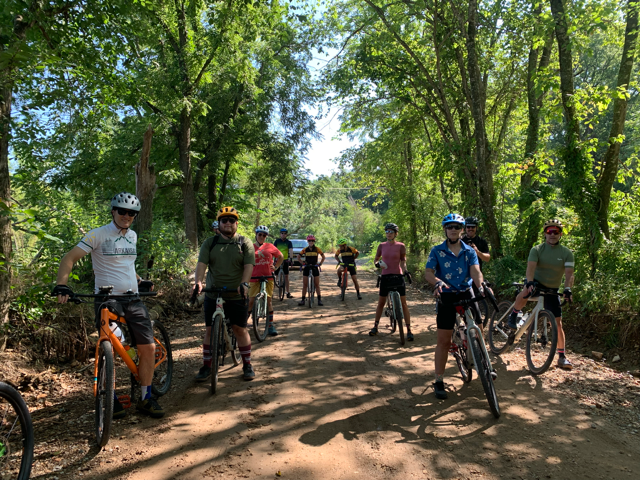 A group of riders pose for a group photo on a gravel road with bright green trees on both sides of the road.